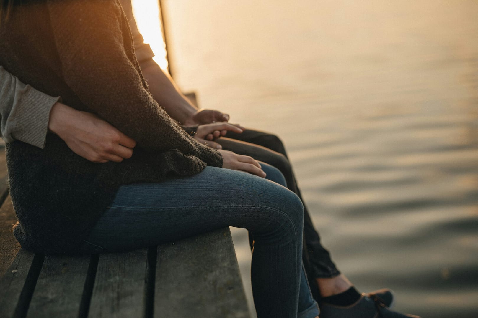 Eine Nahaufnahme von zwei Personen, die auf einem Steg sitzen. Im Mittelpunkt stehen ihre gefalteten Hände, die Vertrauen symbolisieren. Die untergehende Sonne spiegelt sich sanft im Wasser im Hintergrund.