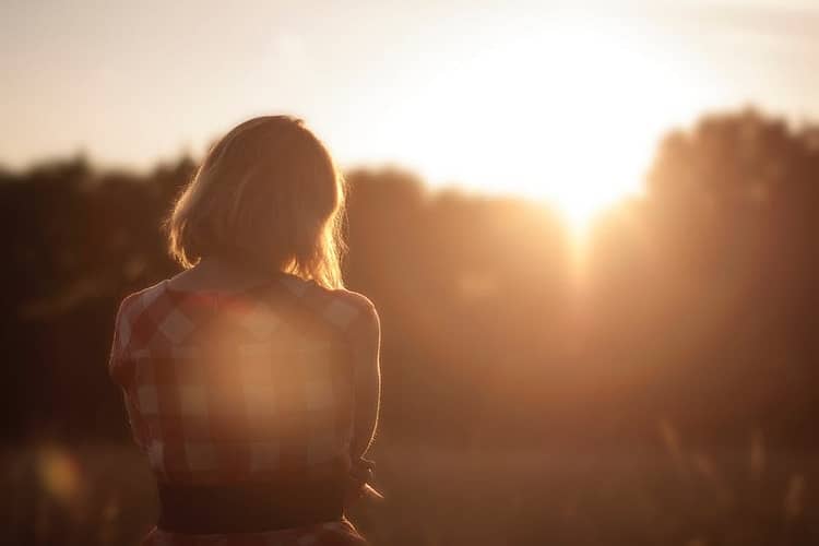 Frau sitzt einsam in der Natur mit dem Rücken zur Kamera und schaut zum Sonnenuntergang.